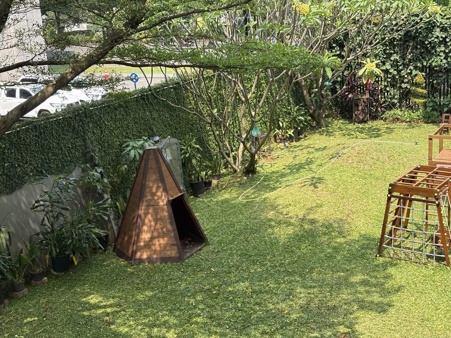 A shaded school garden with a wooden teepee, climbing frame and potted plants, with a city road and parked cars visible just beyond the hedge
