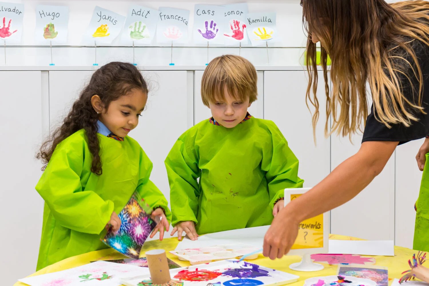 Two young children in green painting aprons working at a table while a teacher guides them, with named handprints displayed on the wall behind