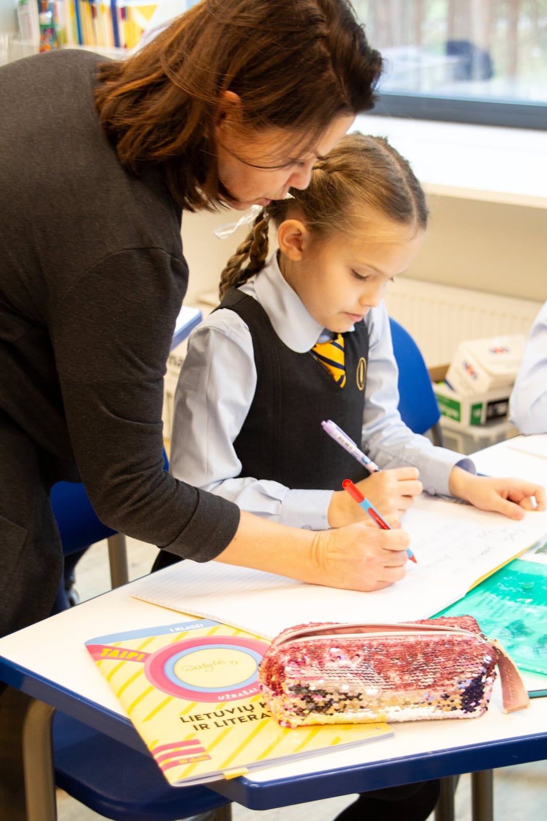 A teacher leaning over a pupil's desk, guiding her handwriting in an exercise book