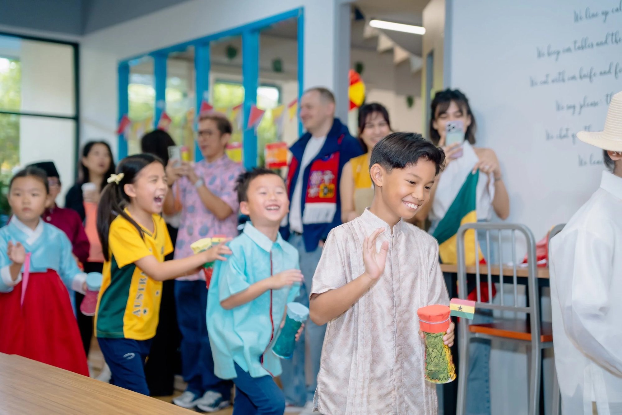 Children in national dress from several countries walking through a classroom carrying handmade instruments and flags while parents watch