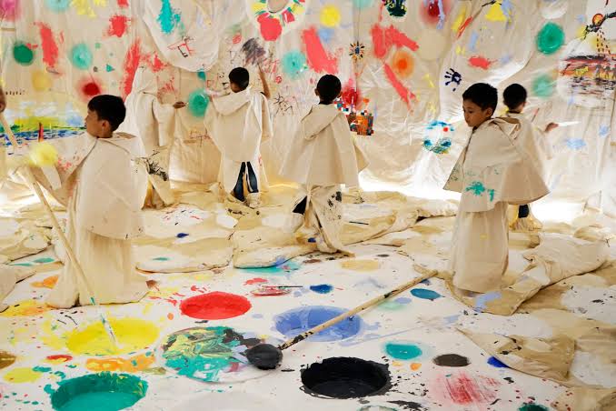 Children in paint-splattered smocks painting on large canvas sheets at an art workshop