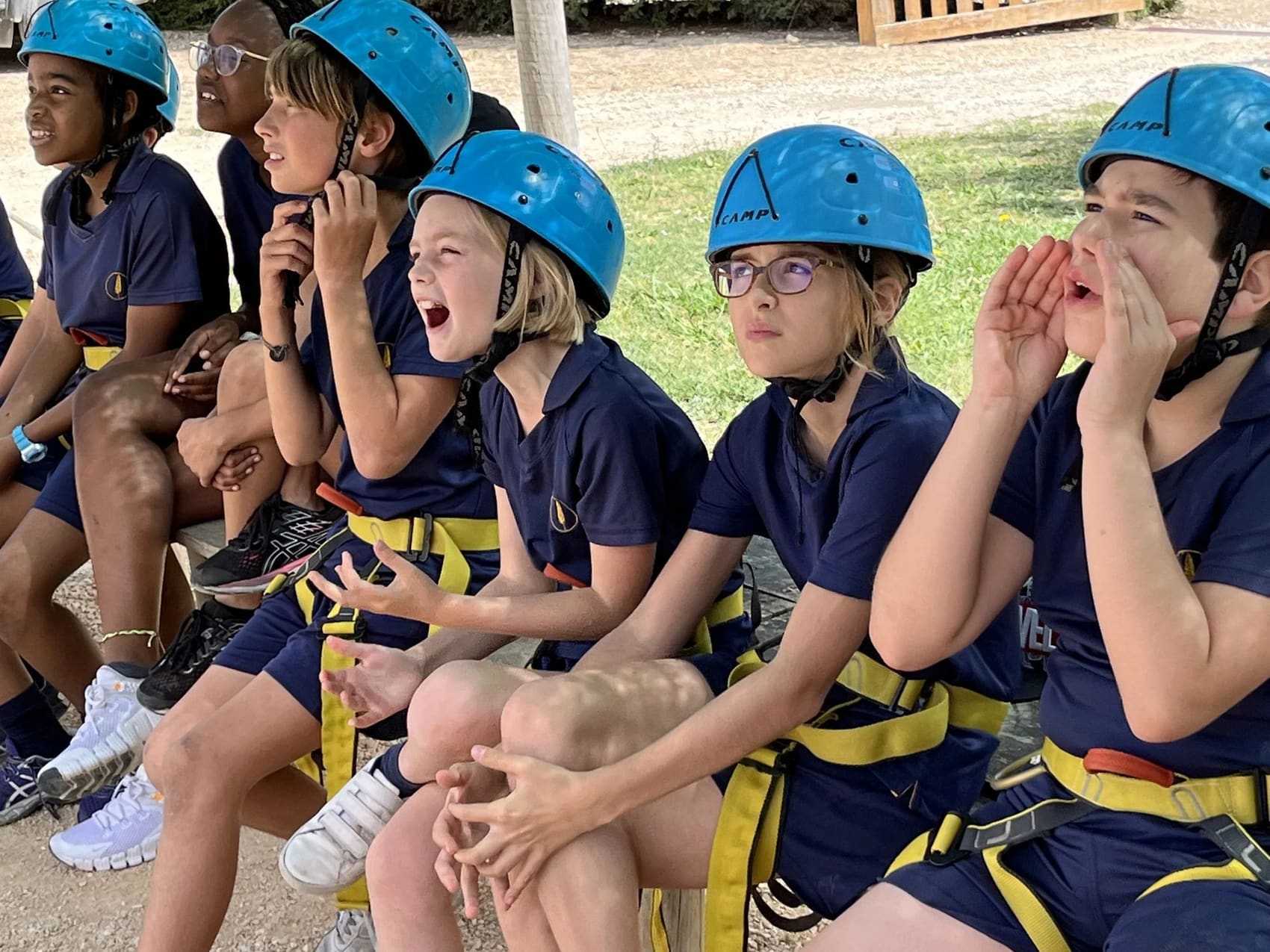 A row of children in climbing helmets and harnesses sitting on a bench outdoors, calling out and cheering towards something off camera