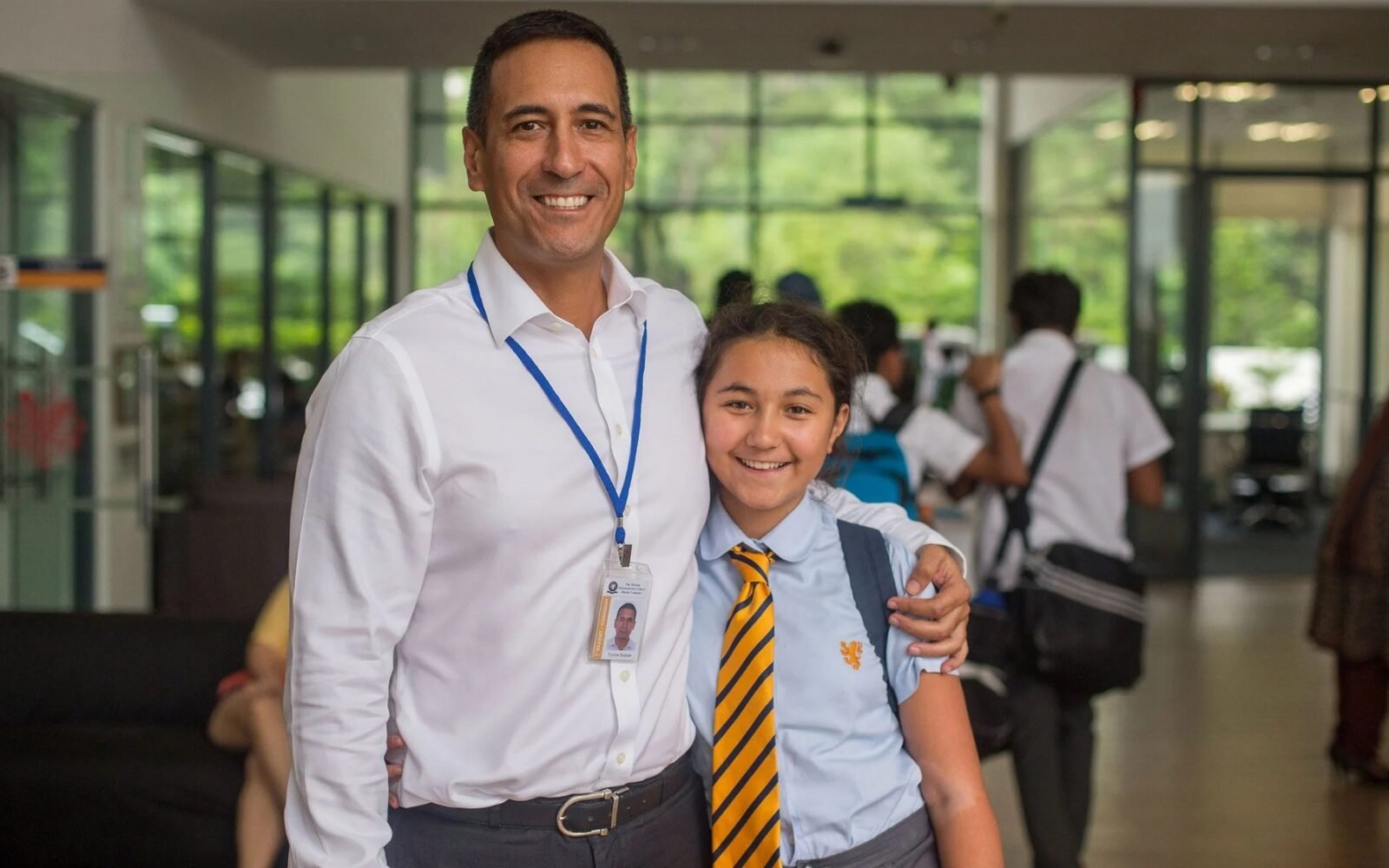 A parent with an arm around their child, who is in school uniform with a backpack, standing in a bright school reception area