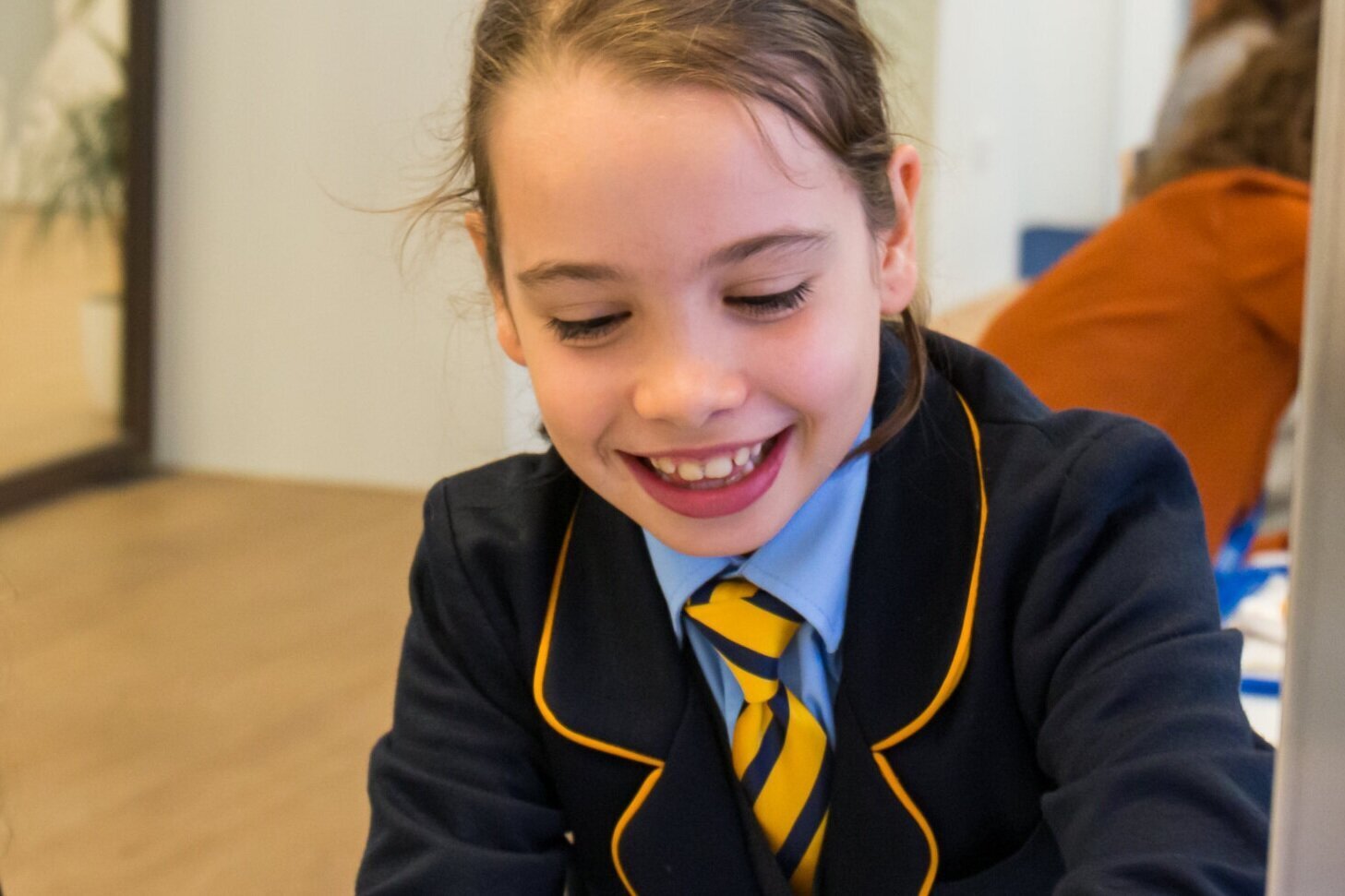 A pupil in a navy blazer and striped tie smiling while working at a desk in a bright classroom