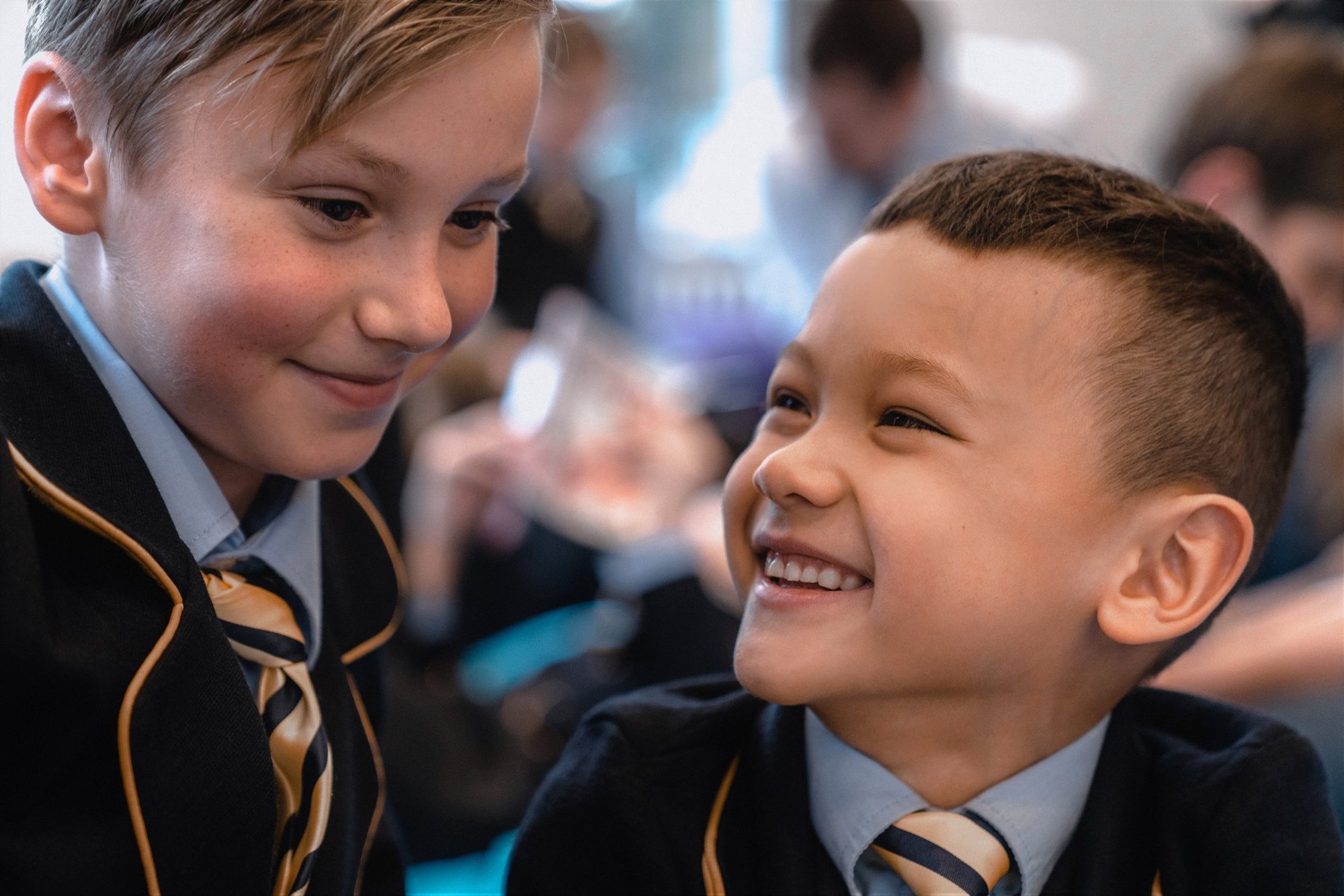 Two boys in school blazers and ties laughing together during a lesson, with classmates out of focus behind them