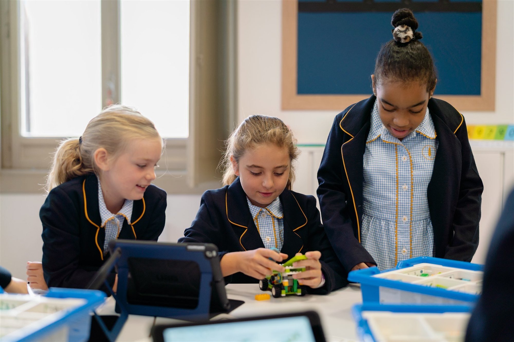 Three pupils in school uniform building a small motorised model together at a classroom table, with tablets and component trays in front of them