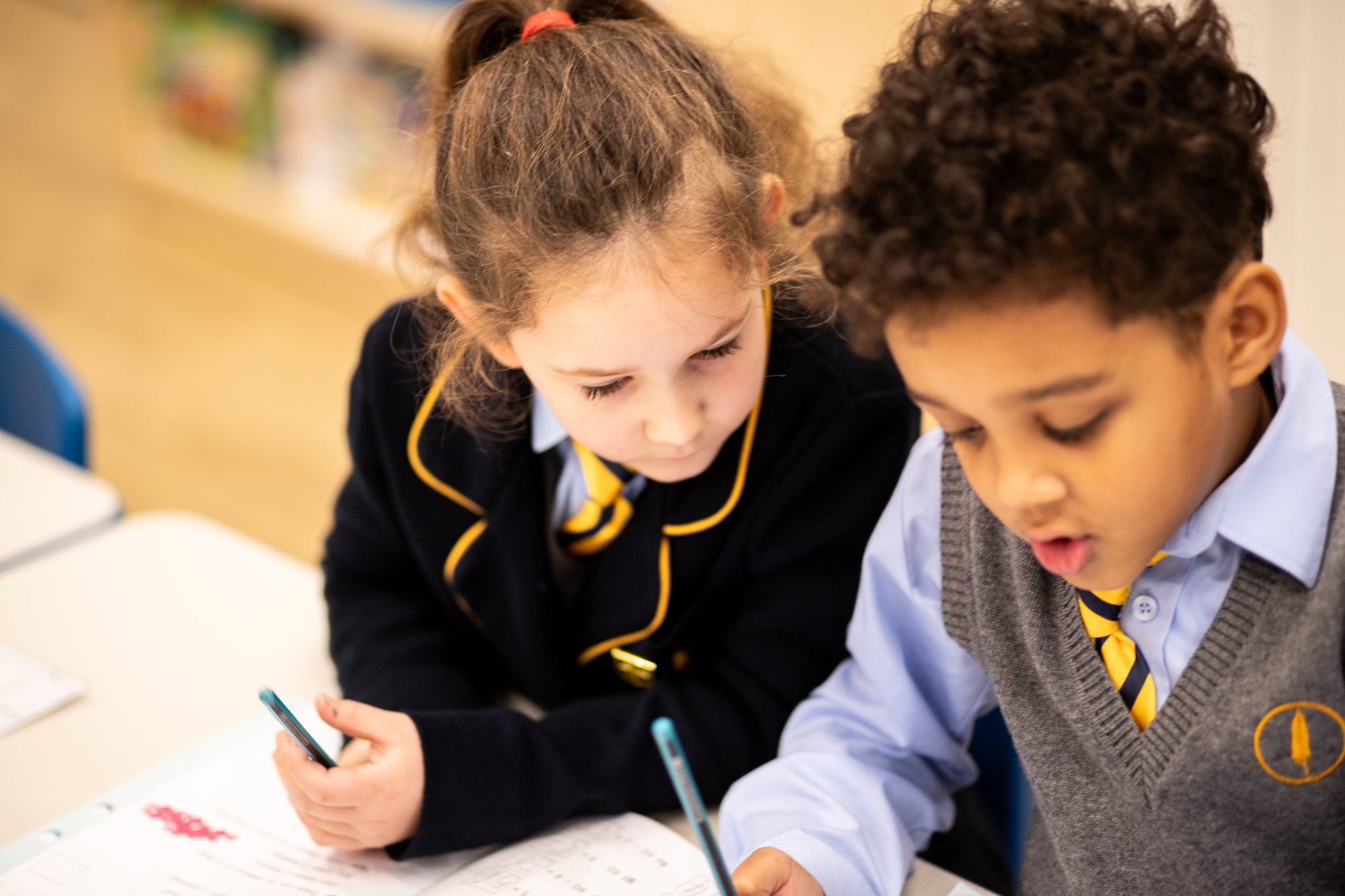 Two young pupils in school uniform working side by side on an exercise book at a classroom desk
