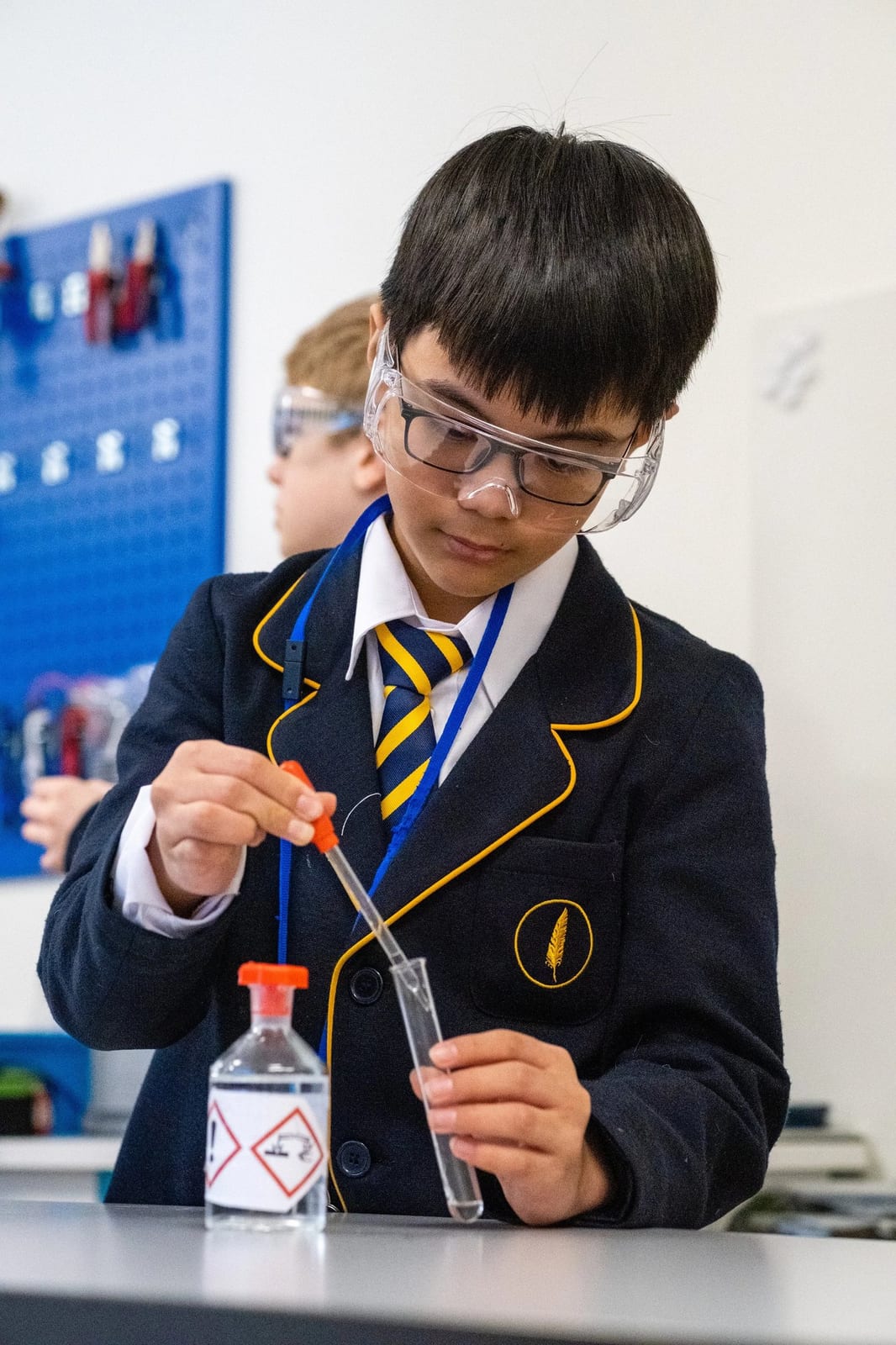 A pupil in blazer and safety goggles drawing liquid into a test tube with a pipette during a science practical
