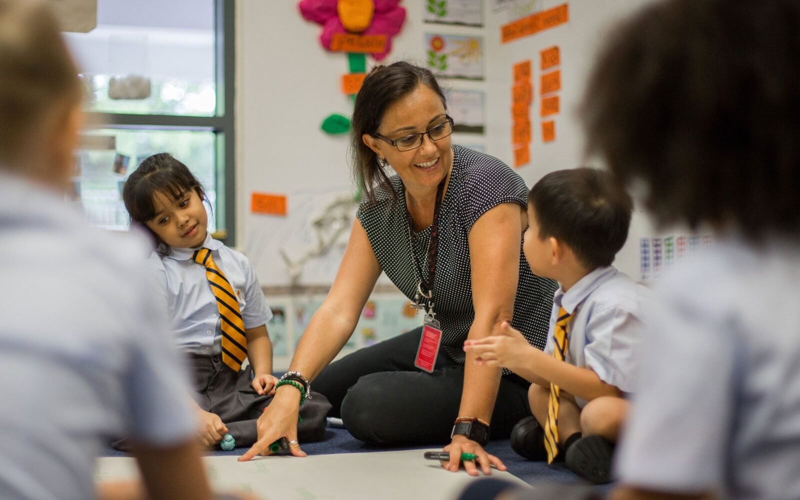 A teacher sitting on the classroom floor with a group of pupils in uniform, working together on a large sheet of paper