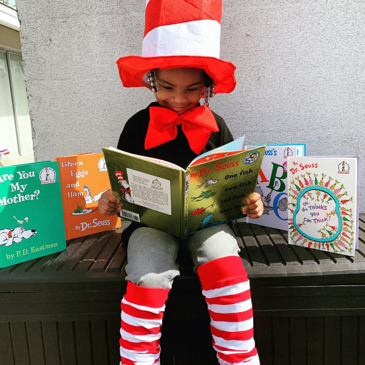 A child in a striped top hat and bow tie reading picture books outdoors on a bench, surrounded by book covers